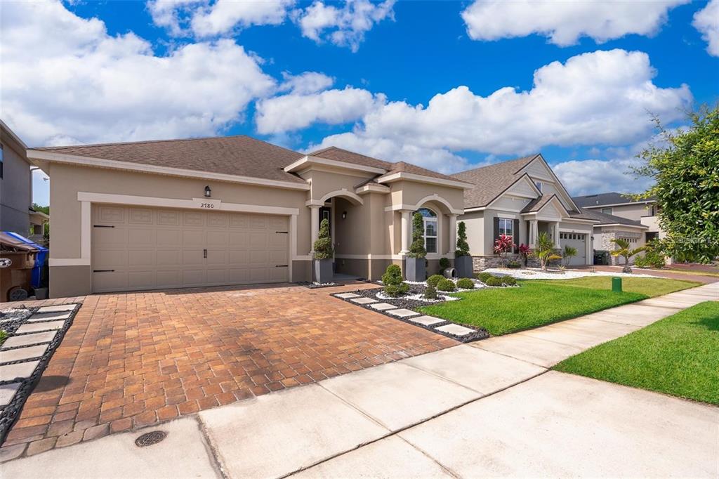 A single-story suburban house with a tan stucco exterior, brick paver driveway, and blue sky — 2780 AVIAN LOOP, KISSIMMEE, FL