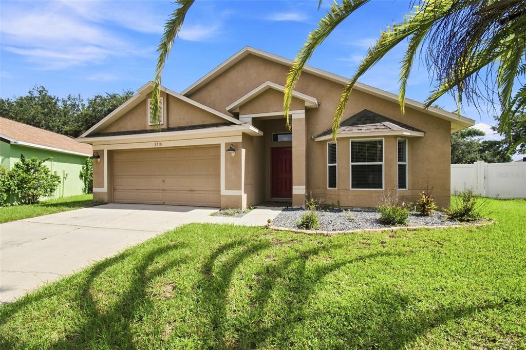 A single-story tan stucco house with a two-car garage, red front door, and front lawn — 9710 WHITE BARN WAY, RIVERVIEW, FL