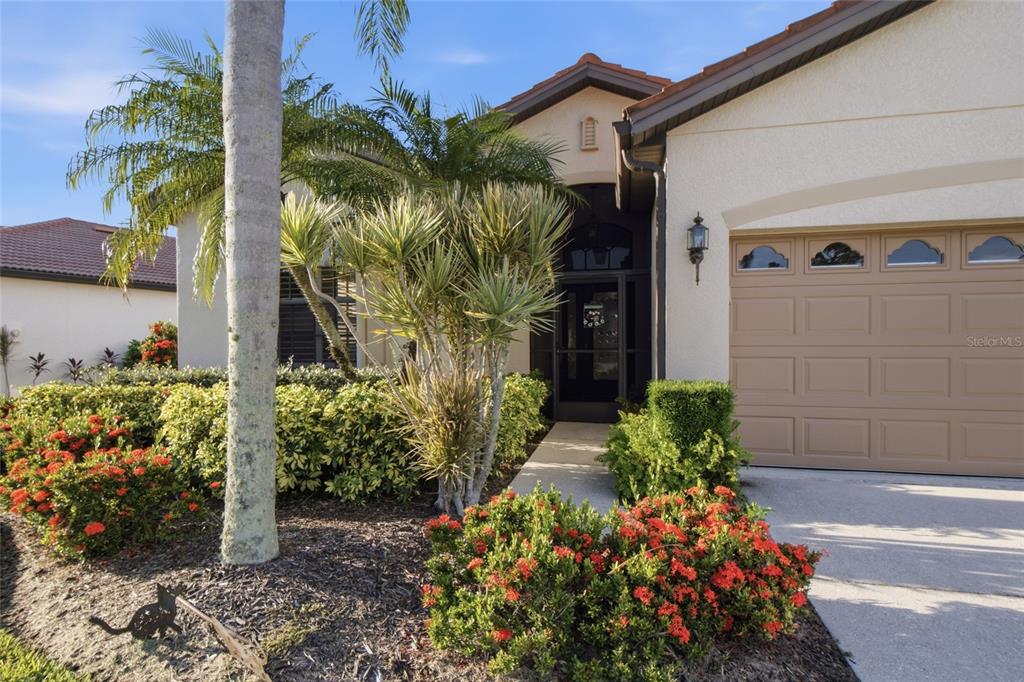 A tan stucco house features a brown garage door, a front entryway, and landscaped garden beds — 1884 BATELLO DRIVE, VENICE, FL