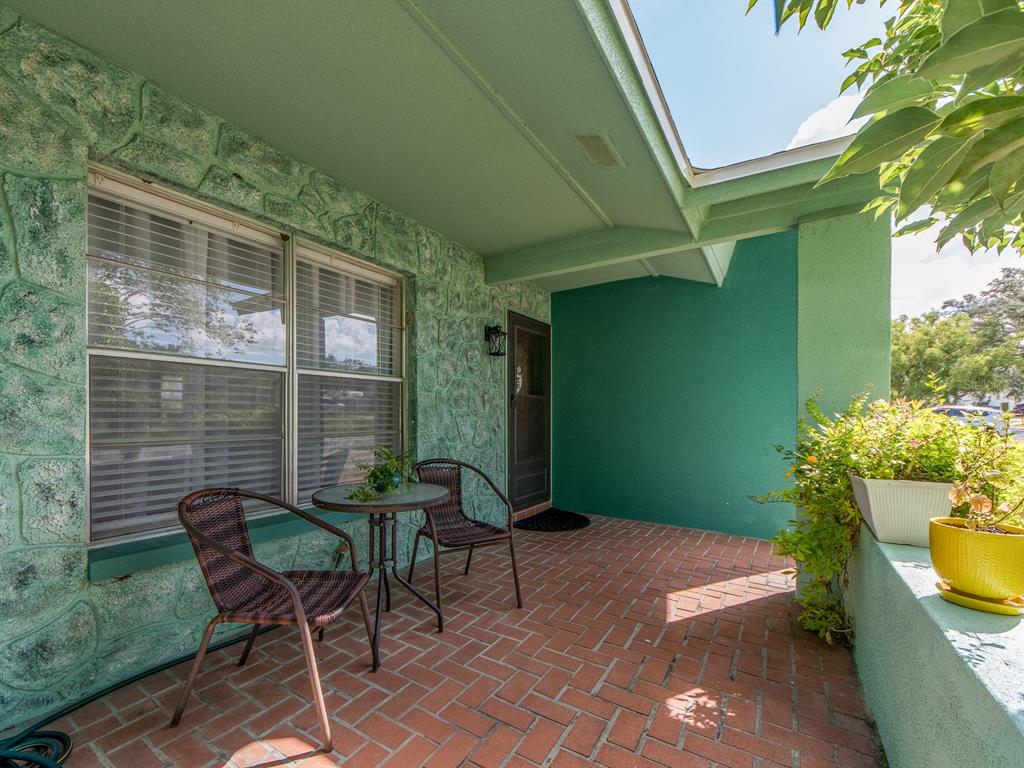 A covered brick porch features a small bistro set and green textured exterior walls — 1832 SETON DRIVE, CLEARWATER, FL