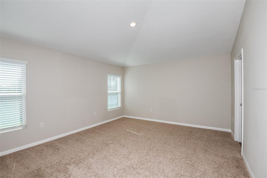 An empty bedroom features beige walls, tan carpeting, two windows, and a vaulted ceiling — 9136 BELLEVIEW FOREST PASS, DADE CITY, FL