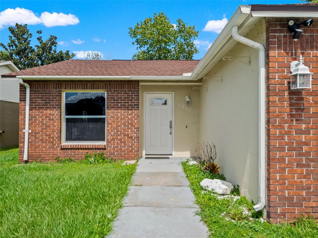 A single-story house exterior features a brick facade, a white front door, and a concrete walkway — 8937 VALENCIA GARDENS DRIVE, ORLANDO, FL