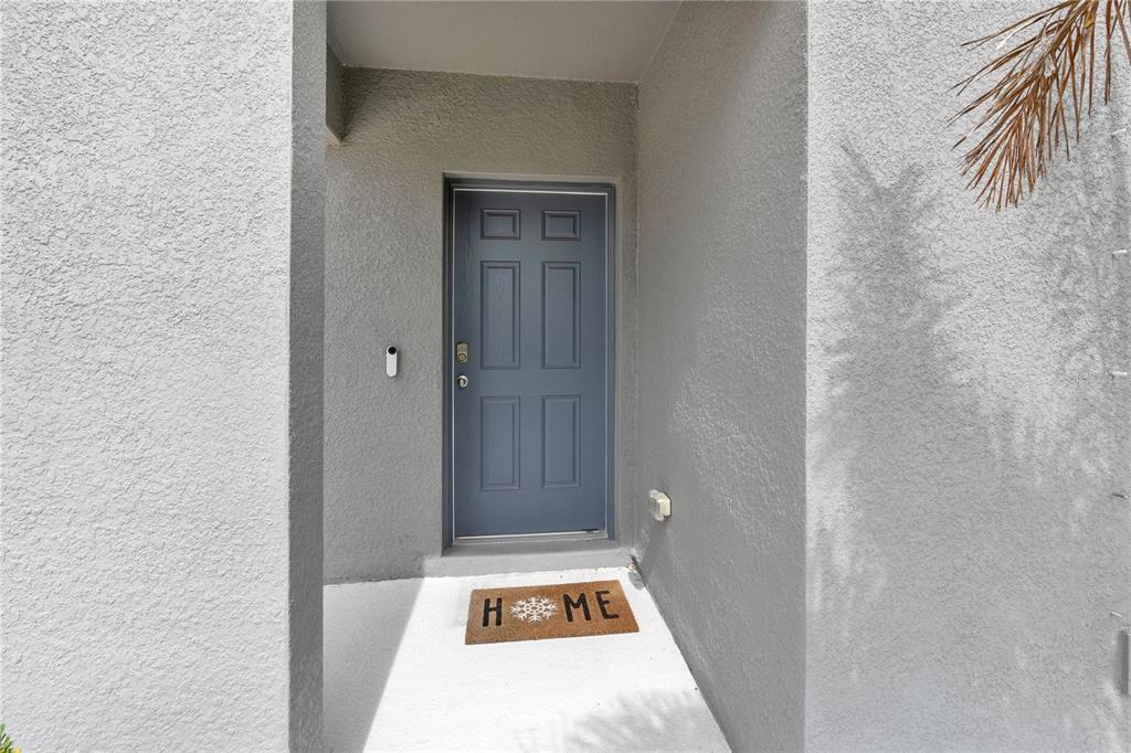 A blue front door with a welcome mat is set within a gray stucco entryway — 12737 TANNENCREST DRIVE, RIVERVIEW, FL