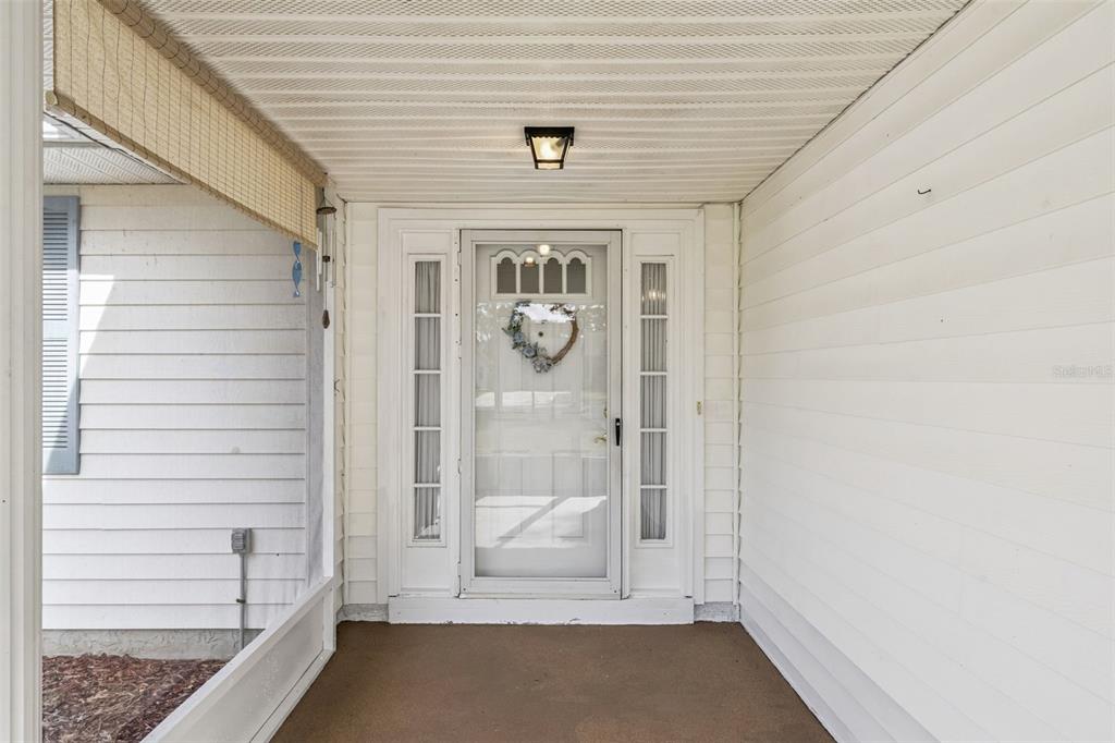 A covered front porch features white siding, a glass storm door, and a concrete floor — 1409 NEW ABBEY AVENUE, LEESBURG, FL