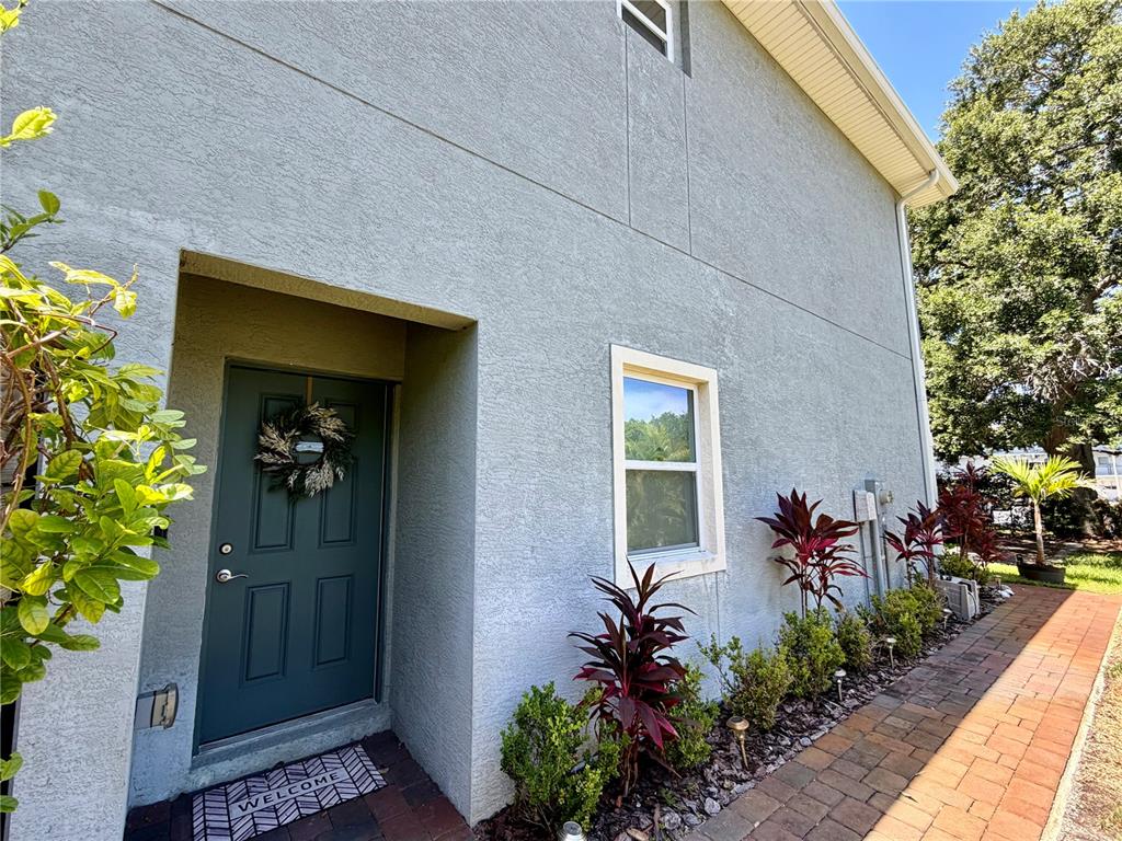 A gray stucco house exterior features a dark green front door and a brick walkway — 281 ASHLEY COURT, DUNEDIN, FL