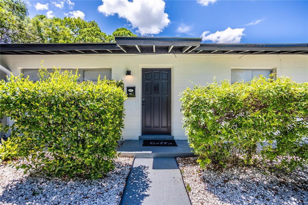 A dark front door is centered between two large green bushes on a gravel yard — 1213 CORBETT LANE, ORLANDO, FL