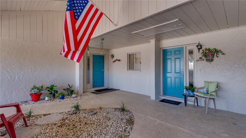 A covered porch features two bright blue doors, a hanging American flag, and potted plants — 1219 16TH CIRCLE SE UNIT 1219, LARGO, FL
