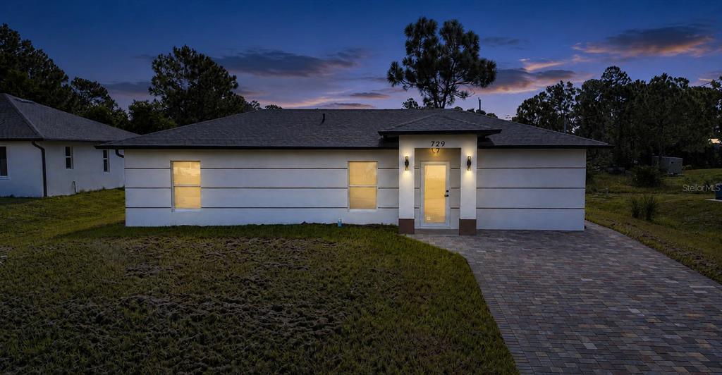 A single-story house with a white exterior, horizontal trim, and a paved driveway at dusk — 729 ANACONDA AVENUE S, LEHIGH ACRES, FL