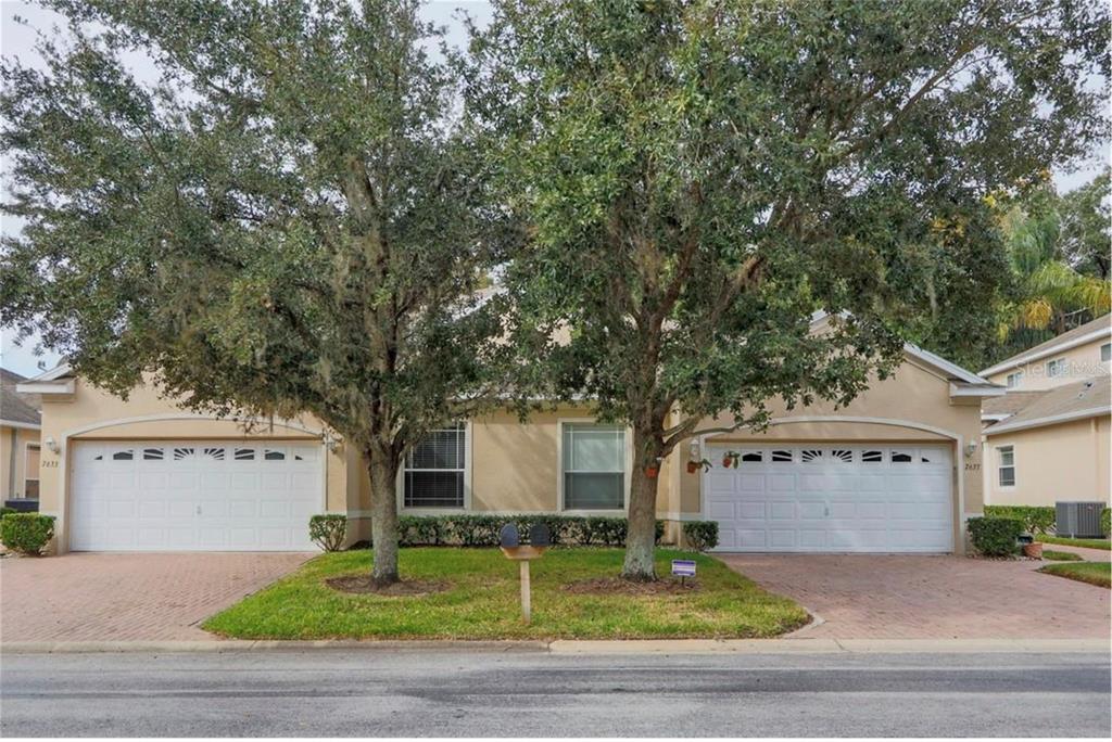 A duplex home with two garage doors, tan stucco walls, and large trees in the front — 7633 WIMPOLE DRIVE, NEW PORT RICHEY, FL