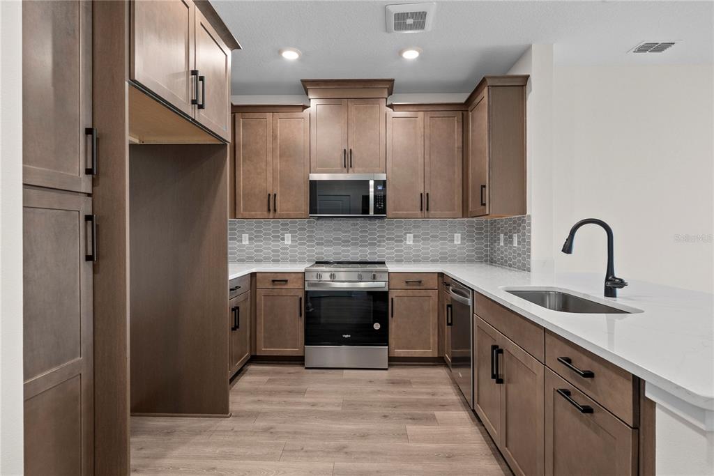 A kitchen features brown cabinets, stainless steel appliances, a gray tile backsplash, and white countertops — 7545 LAUREATE BOULEVARD UNIT 7404, ORLANDO, FL