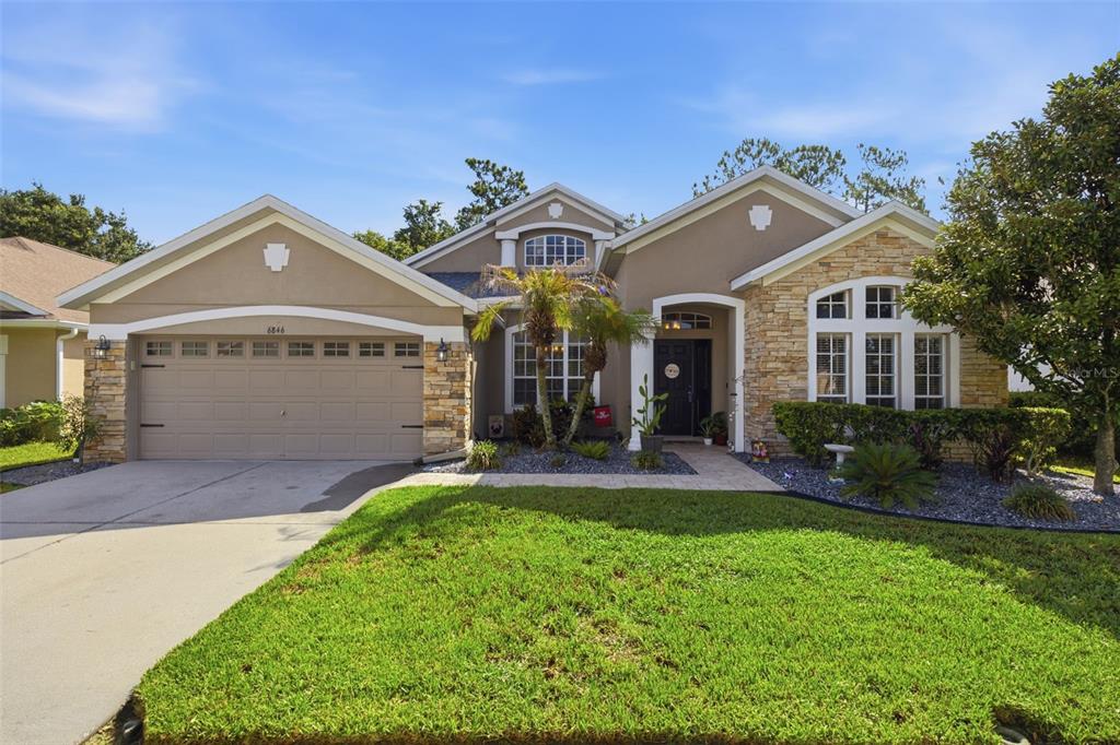 A single-story suburban house with a tan stucco exterior, stone accents, and a two-car garage — 6846 PINE SPRINGS, WESLEY CHAPEL, FL