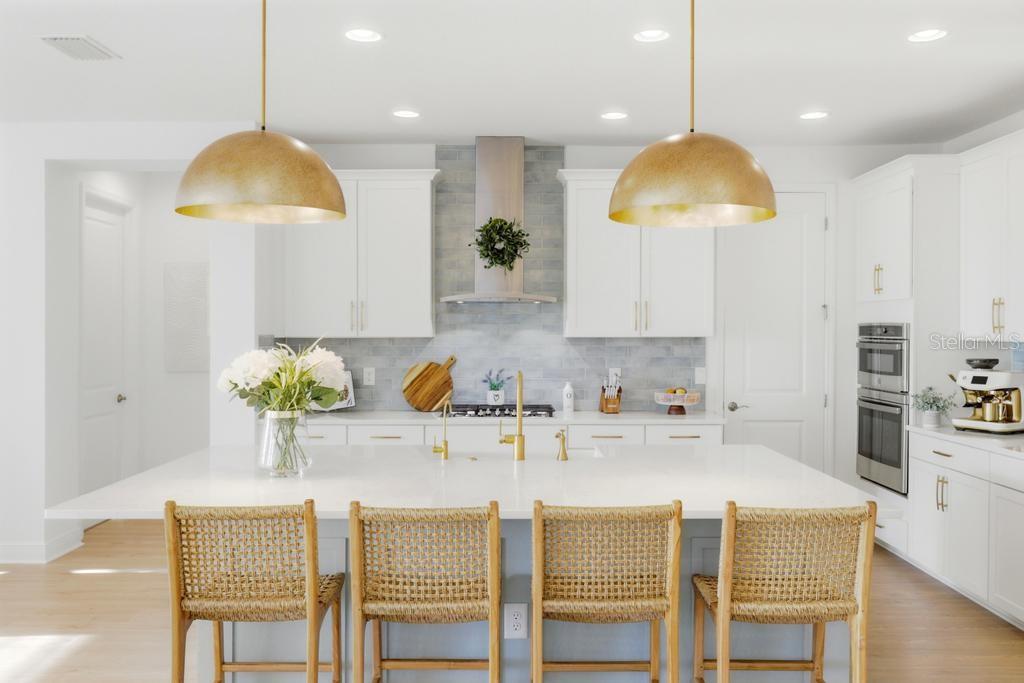 A bright kitchen features a white island with woven stools, gold pendants, and blue backsplash — 17731 SAVORY MIST CIRCLE, BRADENTON, FL