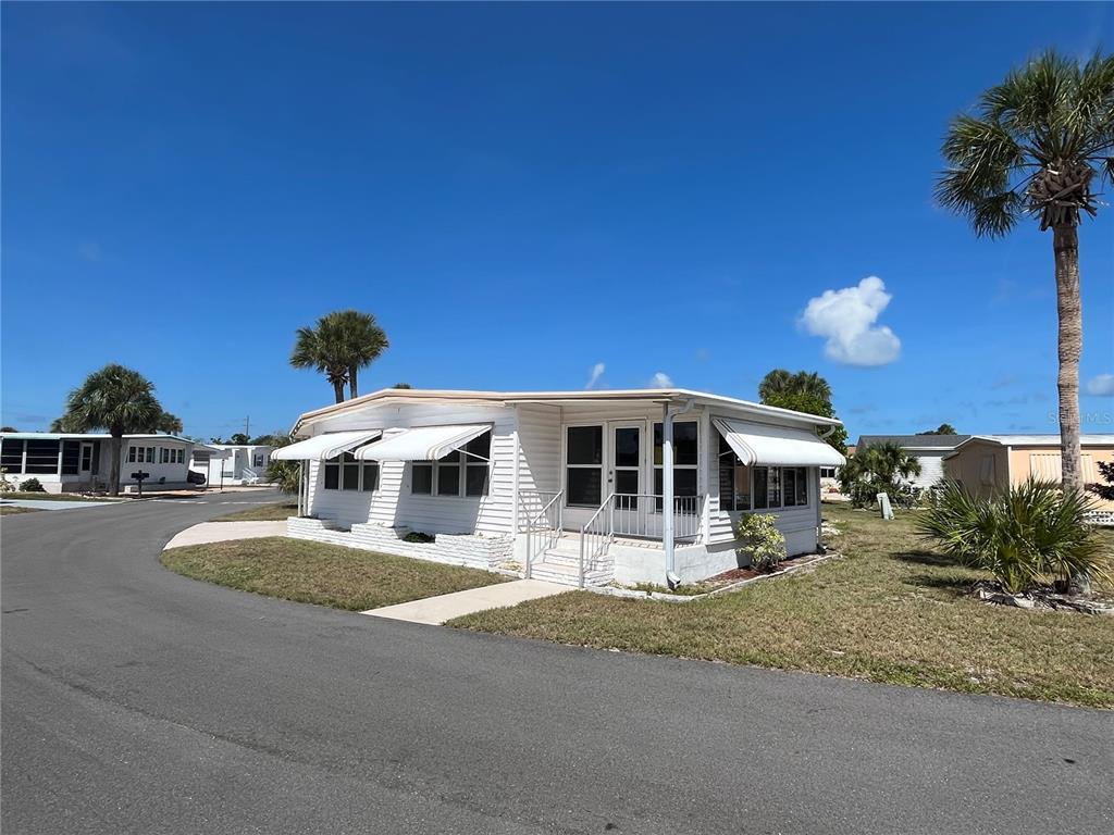 A white mobile home with striped awnings sits on a corner lot under a blue sky — 633 COMO DRIVE, VENICE, FL