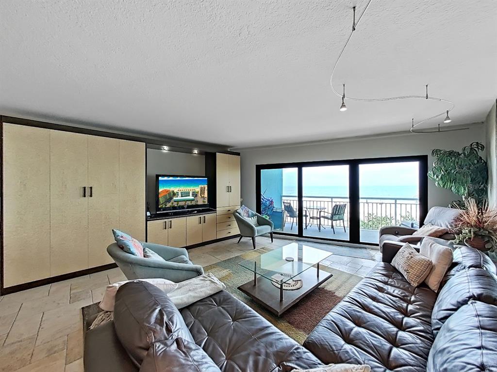 A living room features a large television, leather sofa, armchairs, and floor-to-ceiling windows overlooking the ocean — 19440 GULF BOULEVARD UNIT 104, INDIAN SHORES, FL