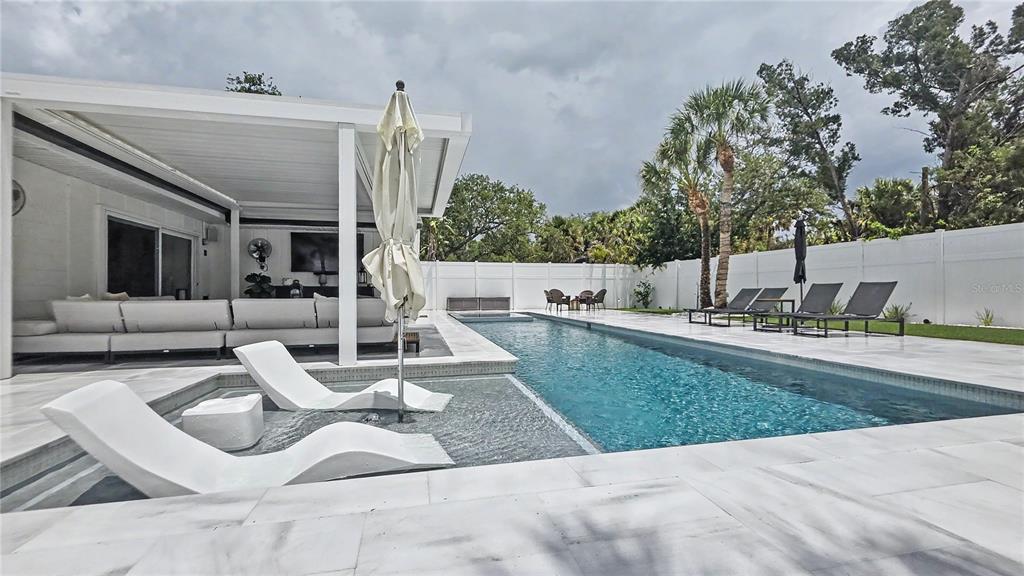 A modern backyard features a rectangular swimming pool with lounge chairs and a covered patio area — 5143 SANDY BEACH AVENUE, SARASOTA, FL