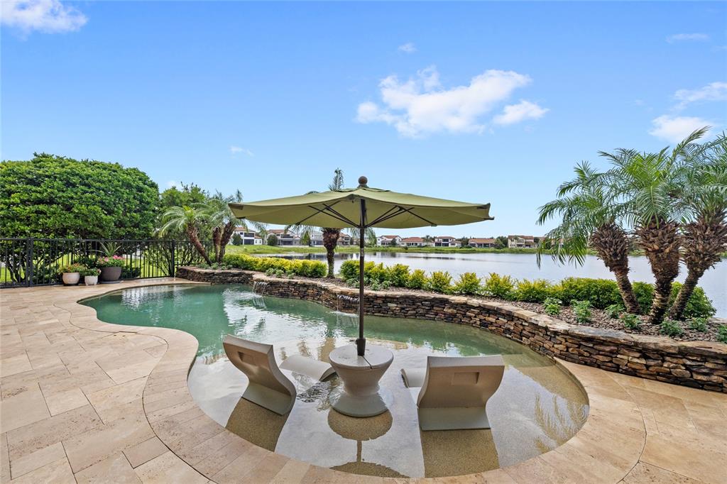 A swimming pool with a tanning ledge, umbrella, and stone wall overlooks a lake — 15475 SHOREBIRD LANE, WINTER GARDEN, FL