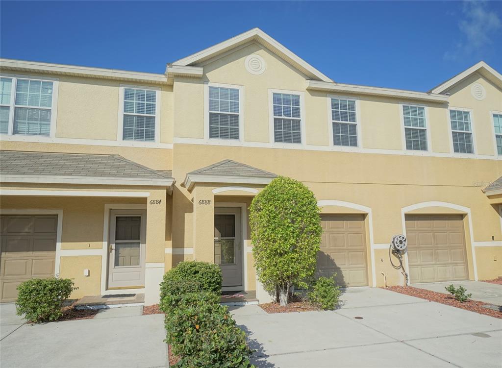 A two-story townhouse exterior features tan stucco walls, a shingled roof, and two garage doors — 6888 46TH LANE N, PINELLAS PARK, FL