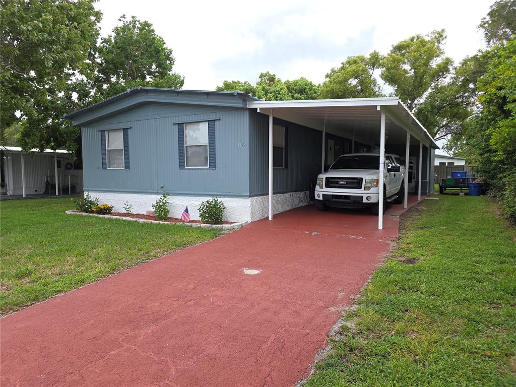 A blue mobile home with a white carport and a red driveway sits on grass — 102 BAREFOOT TRAIL, PORT ORANGE, FL