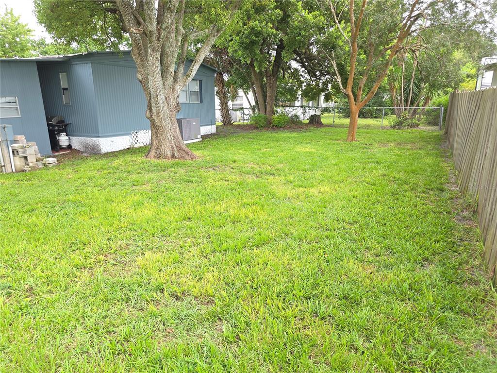 A grassy backyard features a blue mobile home, mature trees, and a wooden privacy fence — 102 BAREFOOT TRAIL, PORT ORANGE, FL