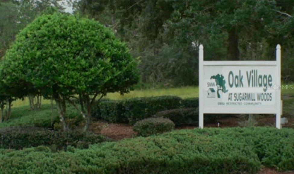 A sign for Oak Village at Sugarmill Woods stands beside a large, manicured green bush — 13 SCABIOSA COURT, HOMOSASSA, FL