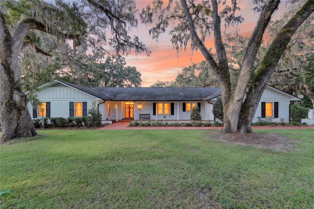 A single-story white house with black shutters sits behind large oak trees at sunset — 1311 SE 17TH AVENUE, OCALA, FL