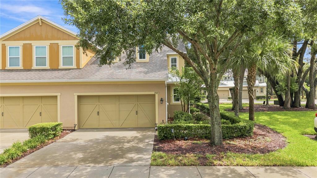 A two-story townhouse exterior with a tan stucco facade, a double garage, and mature trees — 17013 VILESTA DRIVE, LUTZ, FL