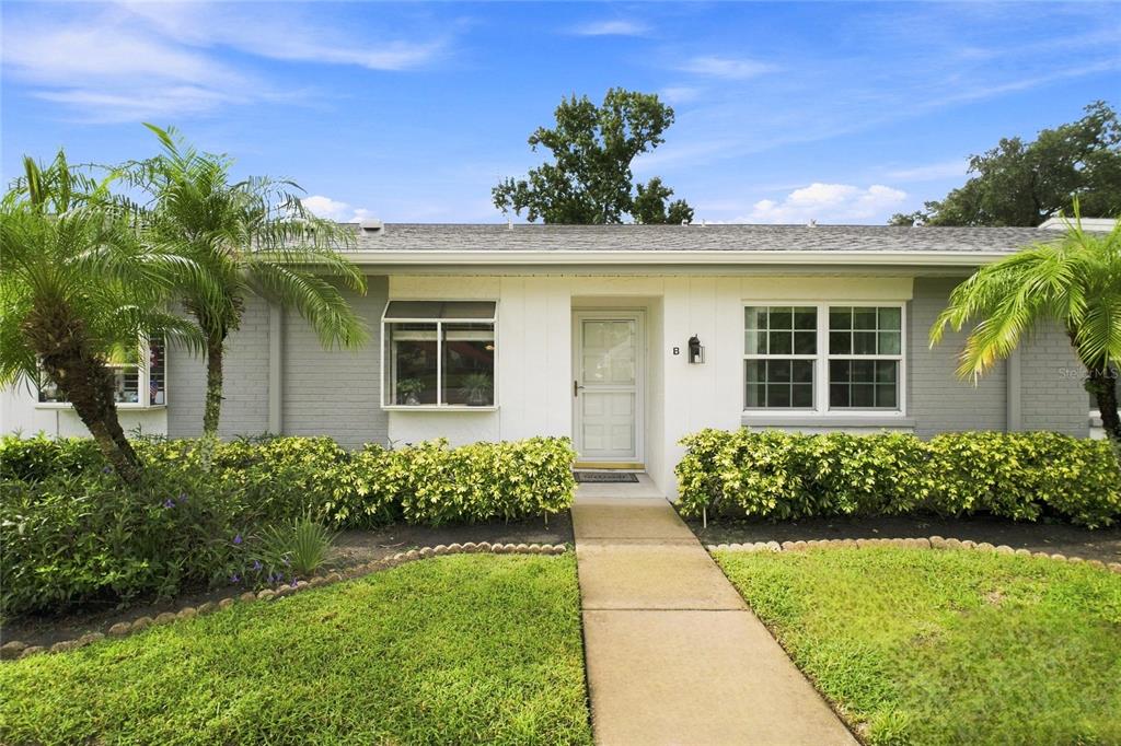 A single-story house exterior features a white front door, gray brick walls, and palm trees — 3370 STONEHAVEN COURT W UNIT B, PALM HARBOR, FL