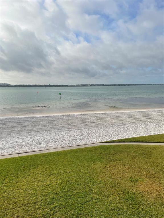 A view of a sandy beach, calm water, and distant shoreline from a grassy lawn — 895 S GULFVIEW BOULEVARD UNIT 303, CLEARWATER BEACH, FL