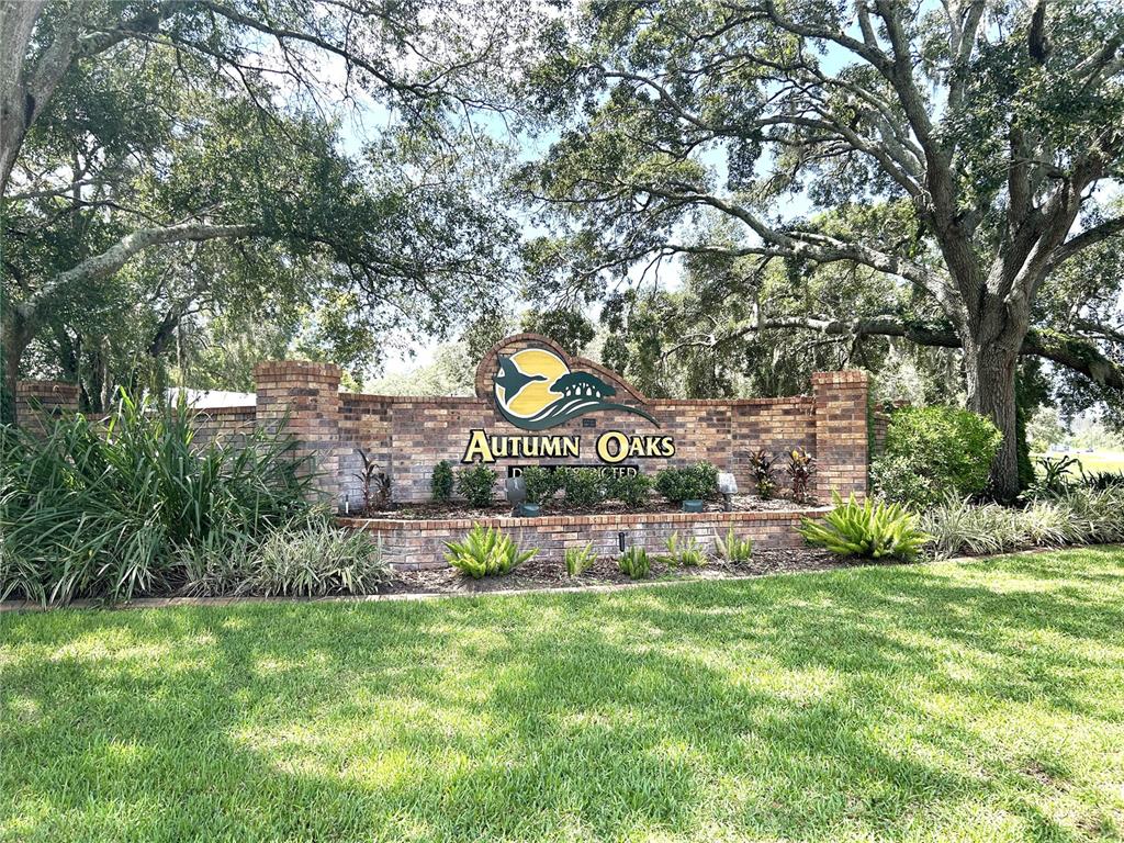A brick entrance sign for Autumn Oaks stands in a grassy area under large trees — 18671 AUTUMN LAKE AVENUE, HUDSON, FL