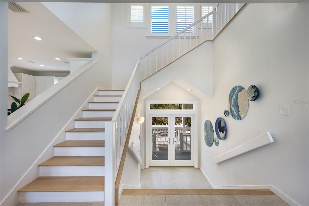 A bright entryway features a wooden staircase, white walls, decorative wall art, and glass doors — 585 BUTTONWOOD BAY DRIVE, BOCA GRANDE, FL