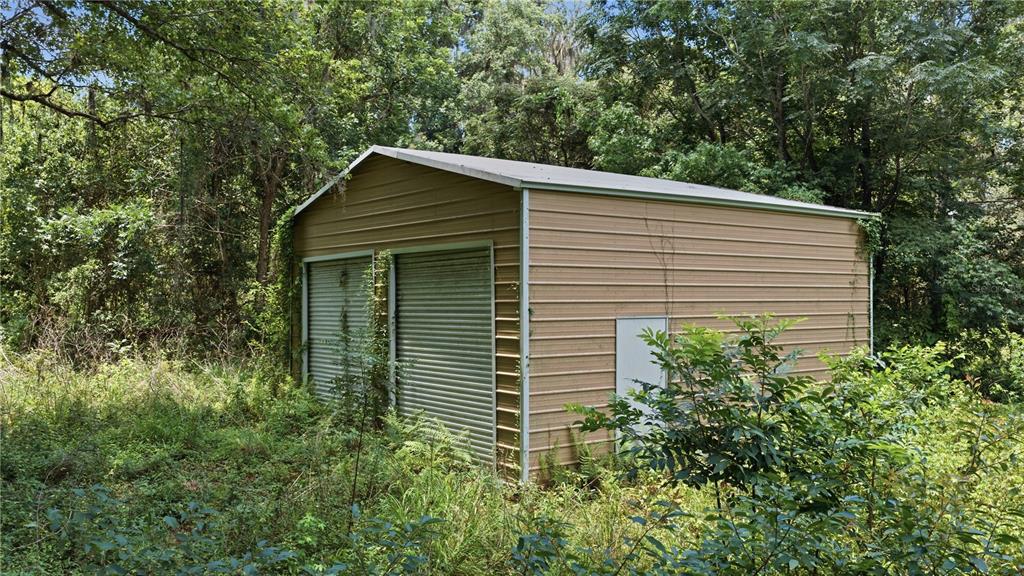 A tan metal storage shed with two roll-up doors sits in a wooded, overgrown area — 17400 NW 120TH AVE ROAD, REDDICK, FL