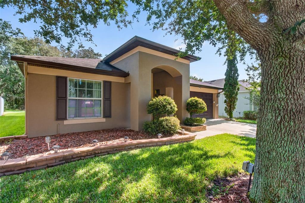 A single-story house with a tan stucco exterior, brown shutters, and a stone-bordered garden bed — 1234 HILL FLOWER DRIVE, BROOKSVILLE, FL