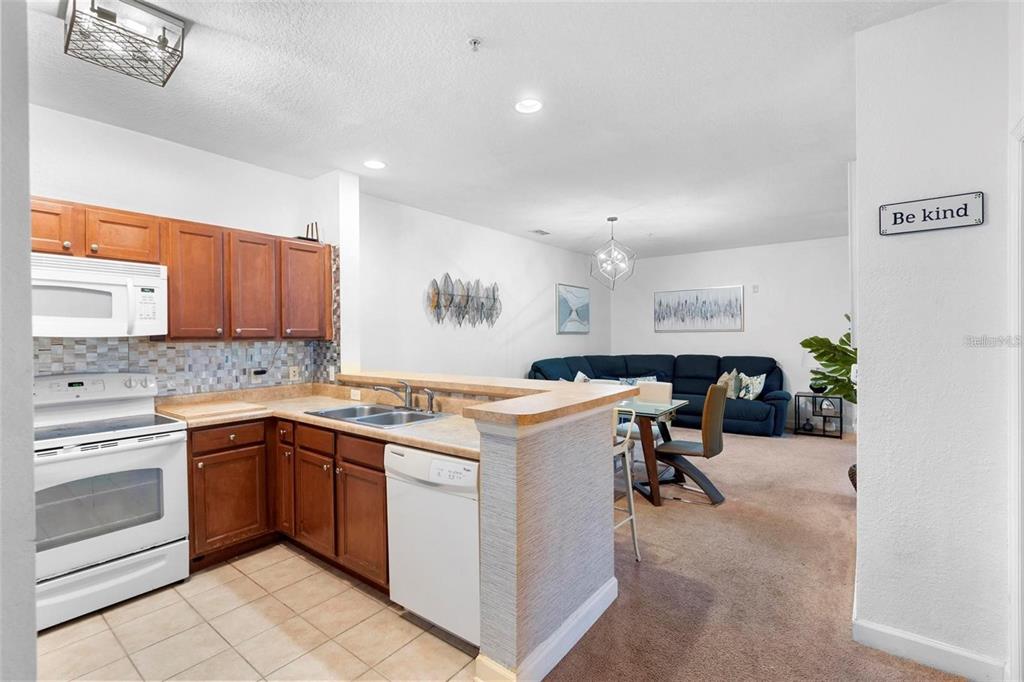 A kitchen with wood cabinets and white appliances opens to a carpeted dining and living area — 3174 FELTRIM PLACE UNIT 203, KISSIMMEE, FL