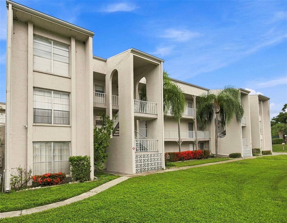 A three-story apartment building with beige stucco walls, balconies, and a green lawn — 2625 STATE ROAD 590 UNIT 1332, CLEARWATER, FL