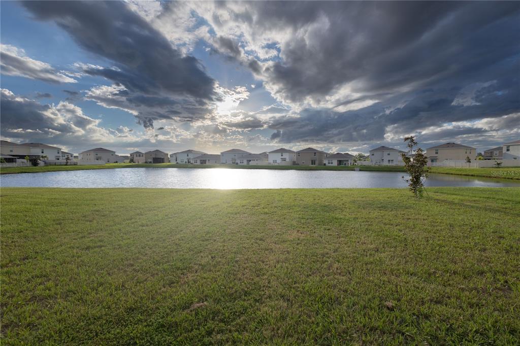A grassy lawn leads to a pond with suburban houses in the background under clouds — 16546 MOSAIC OAR DRIVE, WIMAUMA, FL
