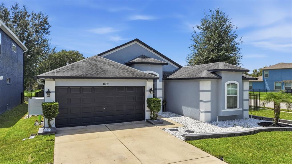 A single-story house with a dark garage door, gray stucco exterior, and a white rock garden — 4037 WARWICK HILLS DRIVE, WESLEY CHAPEL, FL