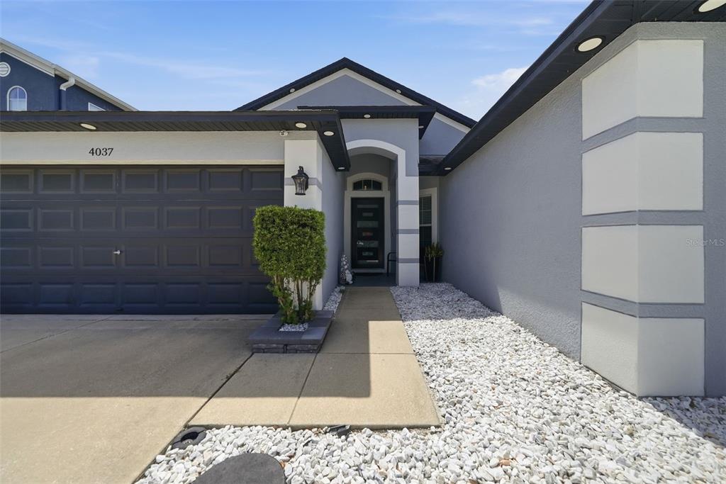 A gray single-story house features a two-car garage, a covered entryway, and white rock landscaping — 4037 WARWICK HILLS DRIVE, WESLEY CHAPEL, FL