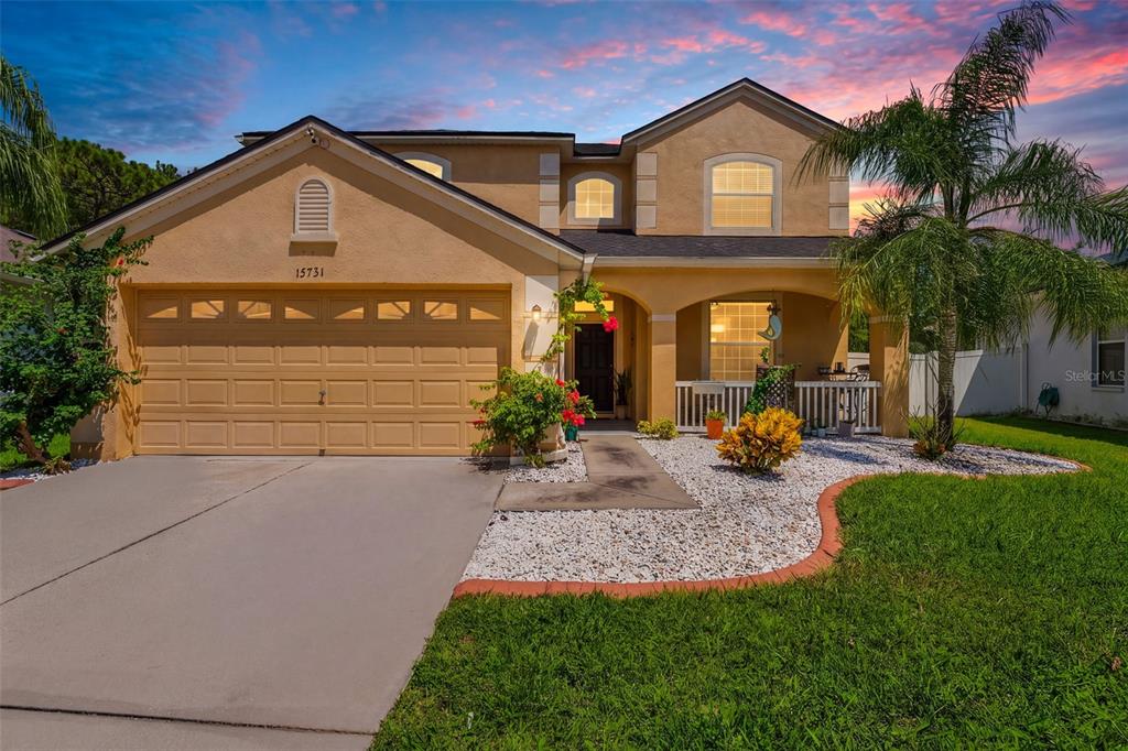 A two-story tan house features a two-car garage, a covered front porch, and landscaping — 15731 CEDAR ELM TERRACE, LAND O LAKES, FL