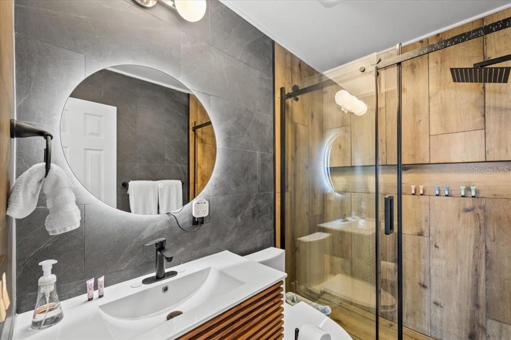 A bathroom features a circular LED mirror, a wood-slatted vanity, and a glass-enclosed shower — 4009 CASA COURT, HERNANDO BEACH, FL
