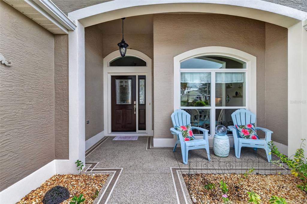 A covered front porch features a dark wood door, two light blue chairs, and gravel — 1473 QUARTER TERRACE, THE VILLAGES, FL