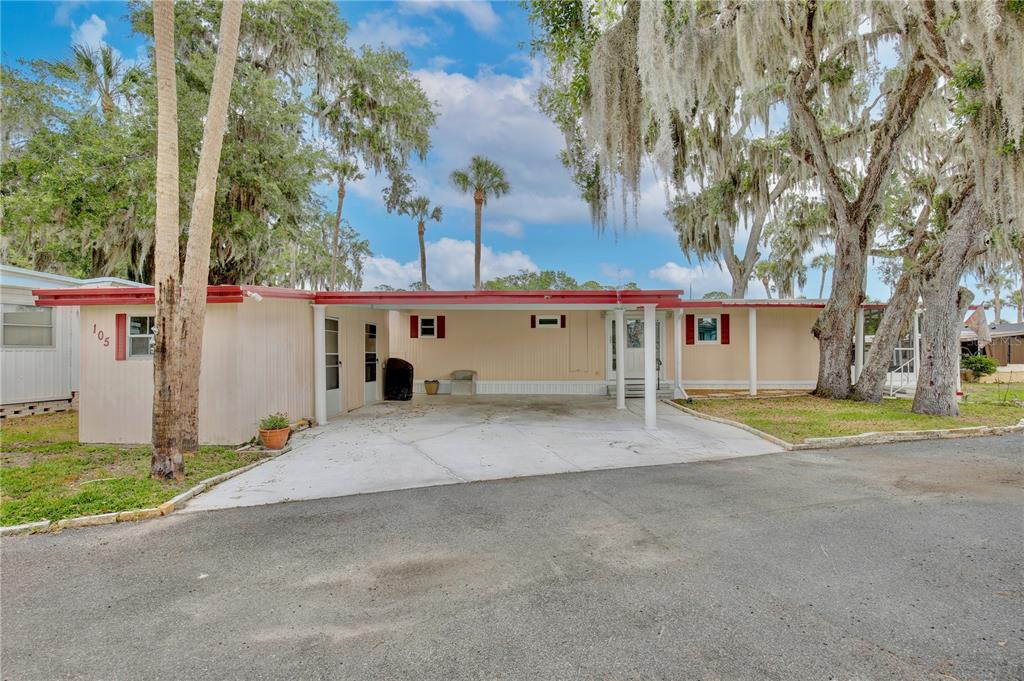 A single-story mobile home with a carport, tan siding, and red trim under mossy trees — 105 CANAL VIEW, EUSTIS, FL