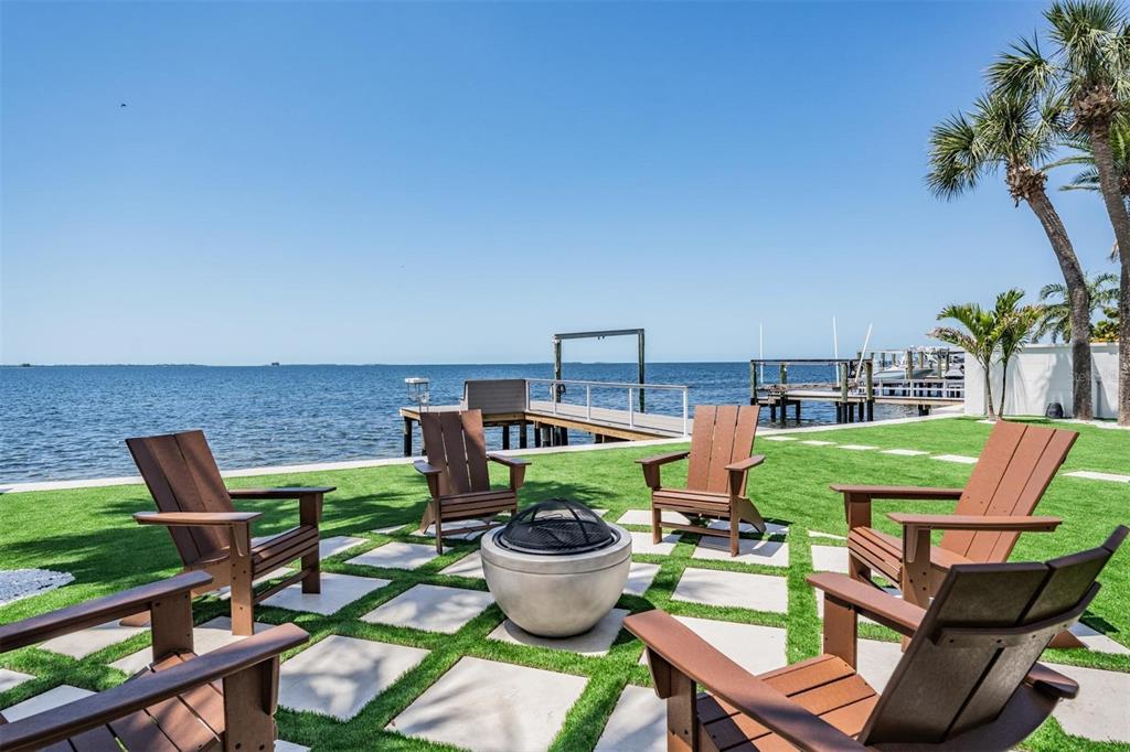 A waterfront backyard features a fire pit, Adirondack chairs, and a dock on a sunny day — 128 CARLYLE DRIVE, PALM HARBOR, FL