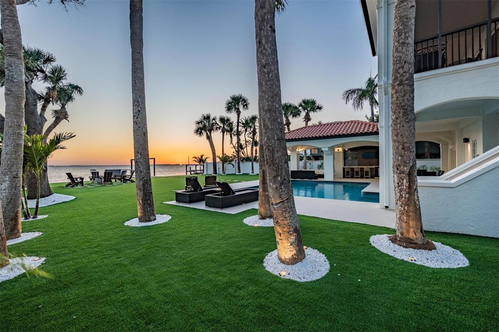 A backyard with a pool, palm trees, and artificial turf overlooking a sunset over water — 128 CARLYLE DRIVE, PALM HARBOR, FL