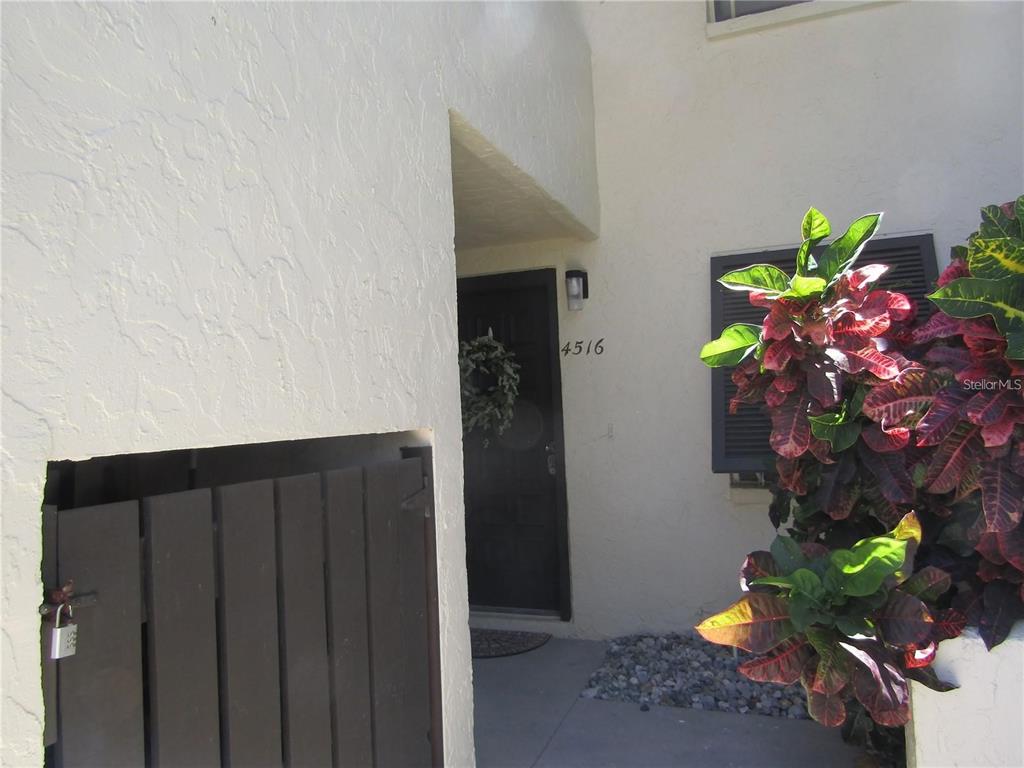A dark front door with a wreath is recessed behind a wooden gate and foliage — 4516 LONGWATER UNIT 29, SARASOTA, FL