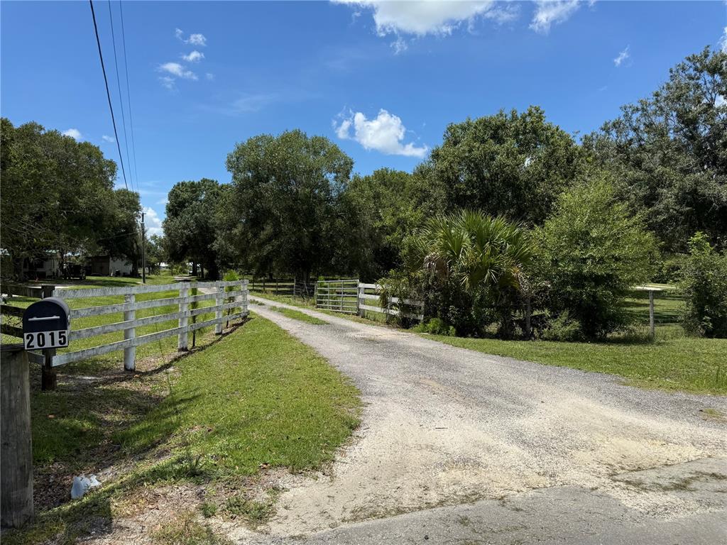 A gravel driveway leads through a white fence toward a wooded area under a blue sky — 2015 NW 46TH AVENUE, OKEECHOBEE, FL