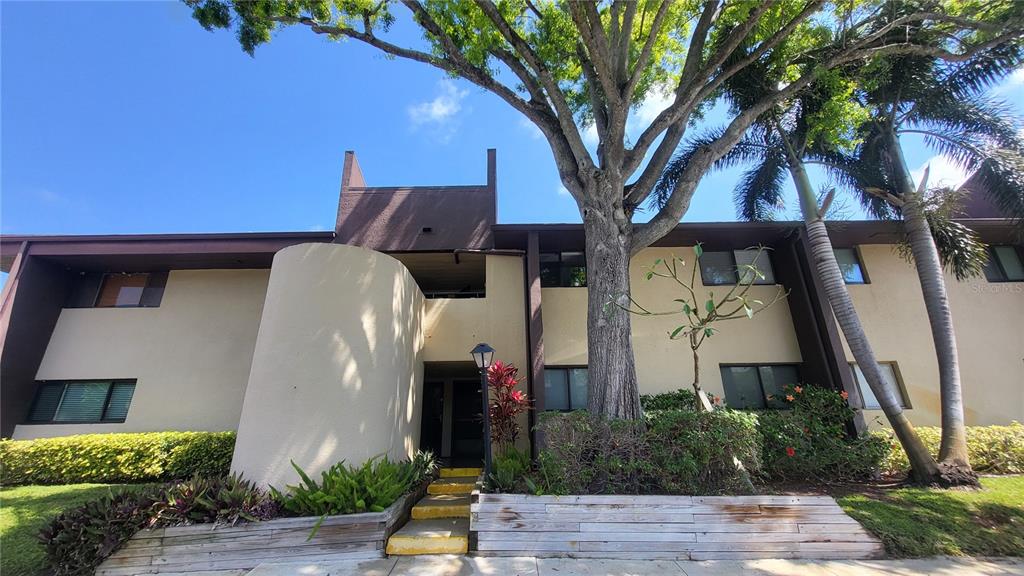 A two-story apartment building exterior features tan stucco walls, dark trim, and a large tree — 875 S VILLAGE DRIVE N UNIT 102, ST PETERSBURG, FL