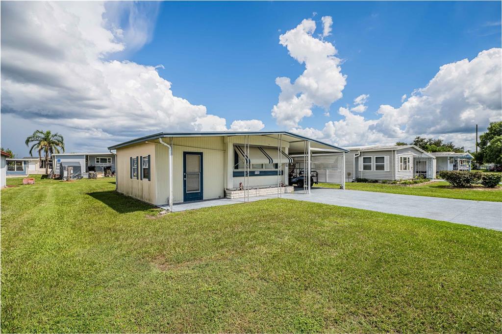 A single-story manufactured home with a carport and a large grassy yard under a blue sky — 5302 POWHATTAN STREET, ZEPHYRHILLS, FL