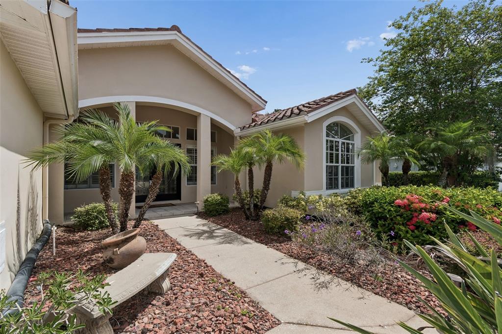 A beige house exterior features a concrete walkway, palm trees, and a stone bench — 1230 BERKSHIRE LANE, TARPON SPRINGS, FL