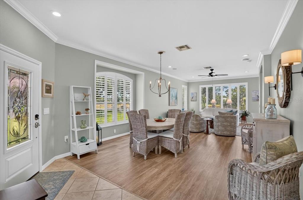 An open-concept dining and living area features light wood flooring, wicker chairs, and plantation shutters — 5037 WHISPERING OAKS DRIVE, NORTH PORT, FL