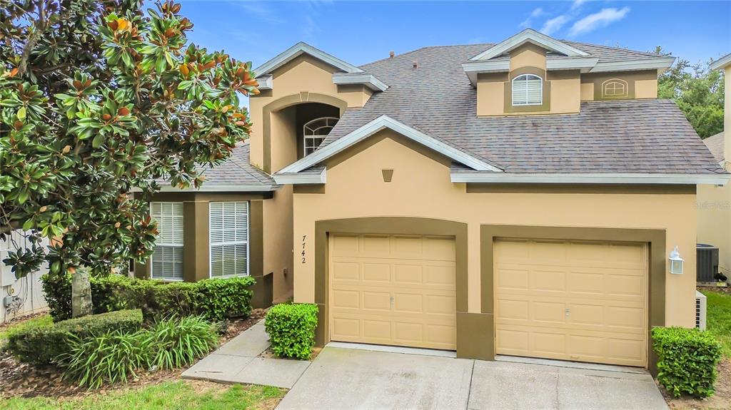 A two-story house with a two-car garage, tan stucco exterior, and a dark shingled roof — 7742 COMROW ST, KISSIMMEE, FL
